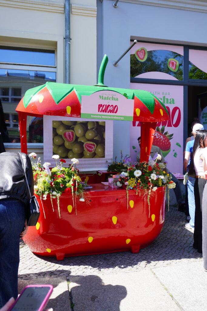 Nucao Yumi Pop UpBunter Verkaufsstand in Erdbeeroptik mit grün-rotem Dach, dekoriert mit frischen Blumen und einem großen Bild von mit Matcha überzogenen Erdbeeren.