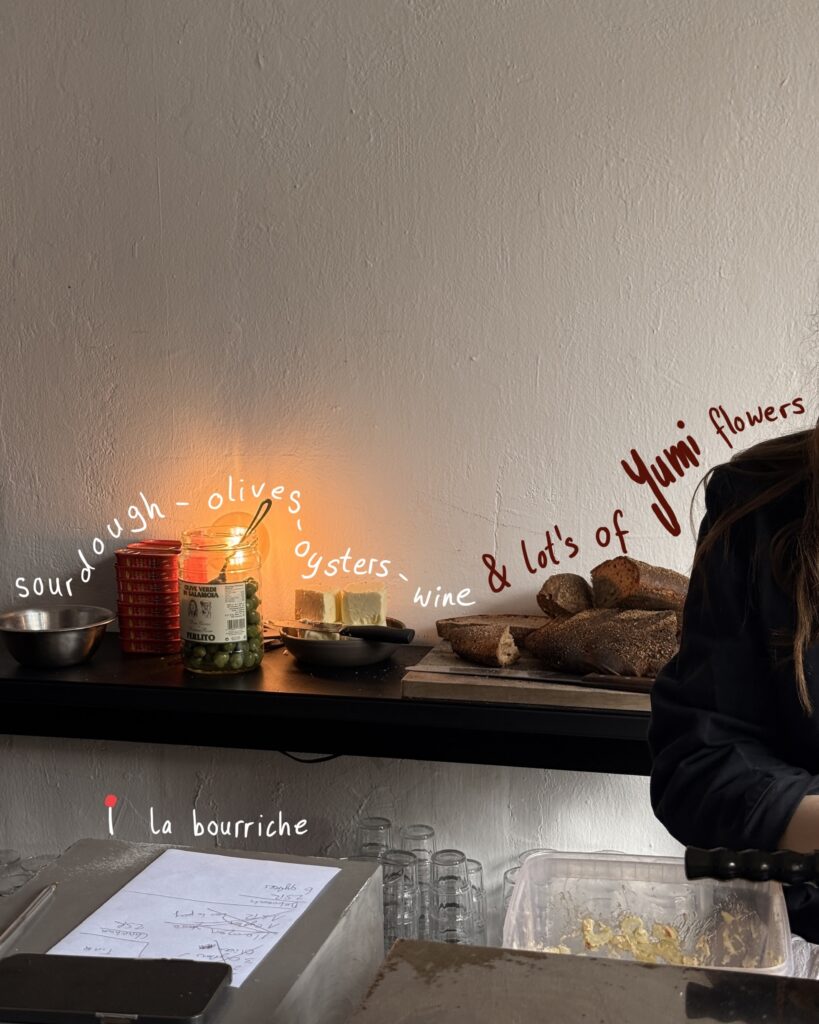 Detailaufnahme einer Theke mit Sauerteigbrot, Butter, Oliven und Wein, dekoriert mit handgeschriebenem Text an der Wand: „sourdough – olives – oysters – wine & lots of Yumi flowers“.