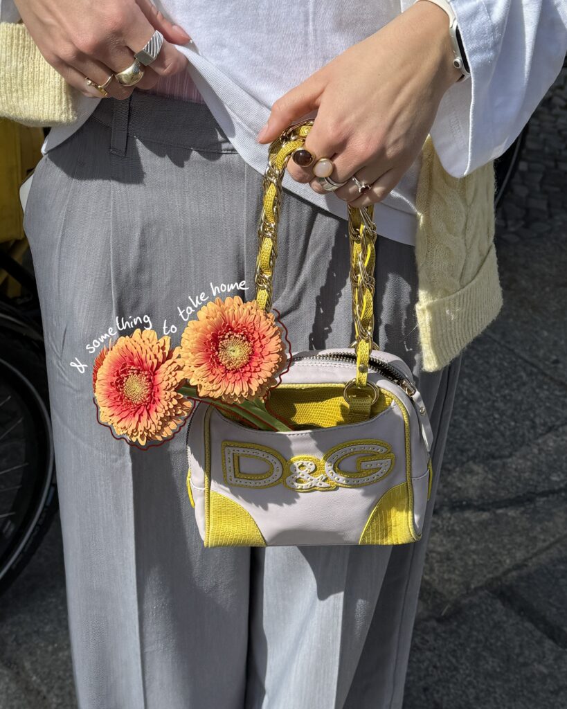 Nahaufnahme einer gelben und weißen D&G-Handtasche, geschmückt mit zwei orangenen Blüten und dem Schriftzug „& something to take home“.dd