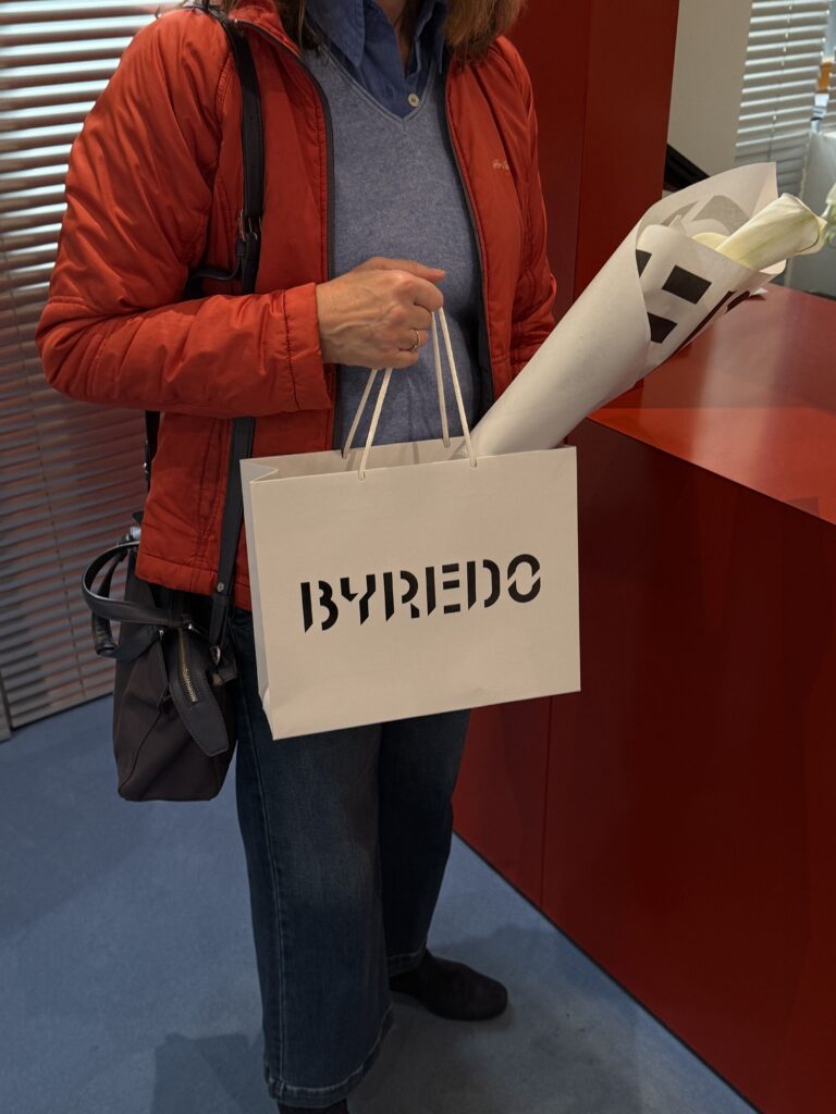 Person in roter Jacke mit BYREDO-Einkaufstasche und einem verpackten Blumenstrauß mit weißen Calla-Lilien im Store.