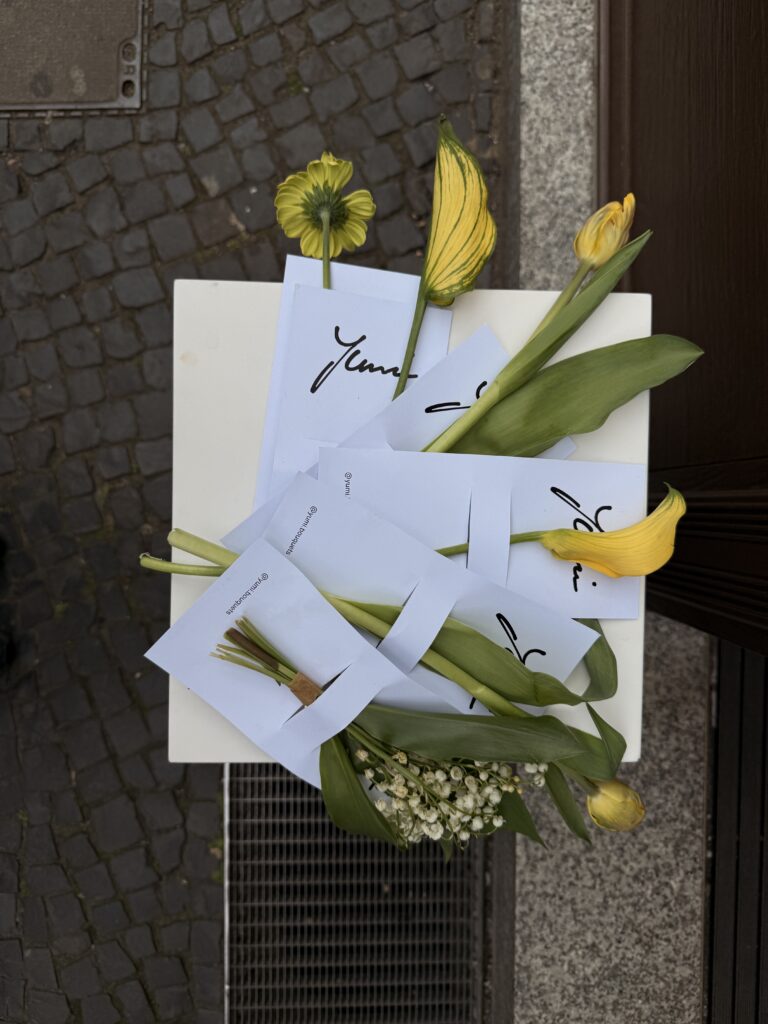  Arrangement aus einzelnen gelben Blumen auf weißen Karten mit Yumi-Logo, platziert auf einem kleinen weißen Tisch auf Kopfsteinpflaster.