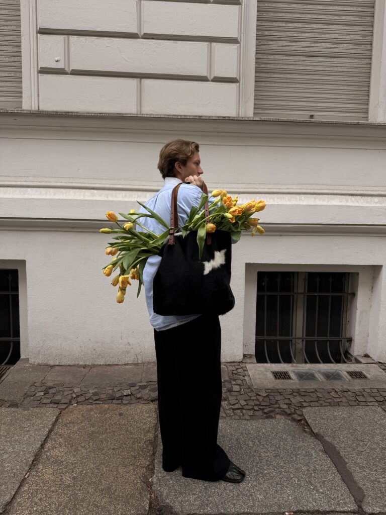Person trägt eine schwarz-weiße Tasche voller gelber Tulpen über der Schulter, steht seitlich auf Gehweg vor einem weißen Gebäude.
