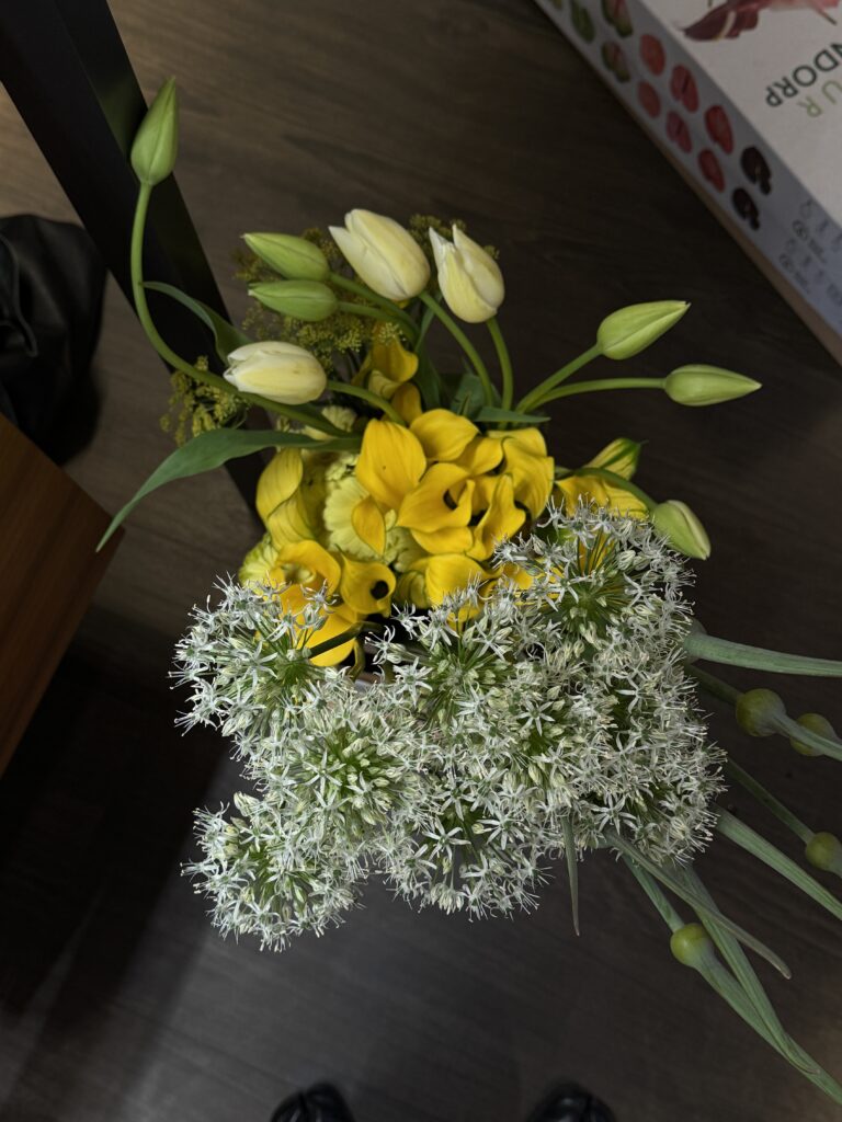 Blick von oben auf ein detailreiches Blumenarrangement mit weißen Blüten, gelben Callas und geschlossenen Tulpenknospen auf dunklem Boden.