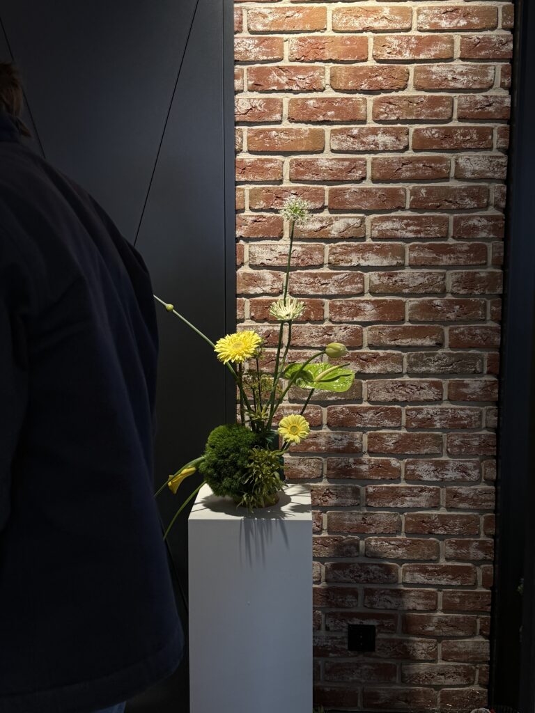 Gelbes Blumengesteck auf weißem Sockel vor einer roten Backsteinwand, teilweise verdeckt durch eine Person in dunkler Kleidung.