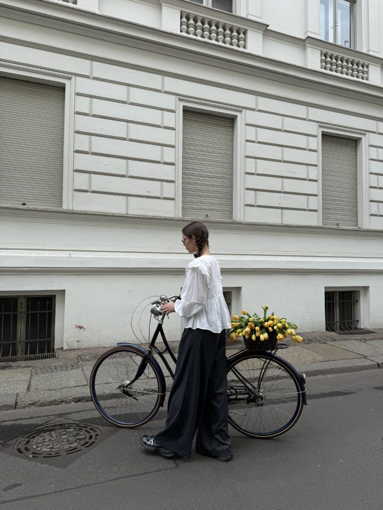 Frau mit weißen Bluse und dunkler Hose steht mit Fahrrad auf der Straße, der Fahrradkorb ist gefüllt mit gelben Tulpen.